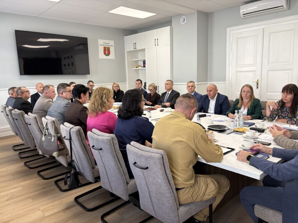 członkowie komisji siedzą przy stole w tle drzwi