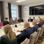 członkowie komisji siedzą przy stole, w tle okna