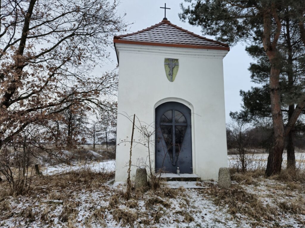 zdjęcie od frontu na kapliczkę, czerwony dach i duże ciemnoszare drzwi, ponad nimi jezus na krzyżu