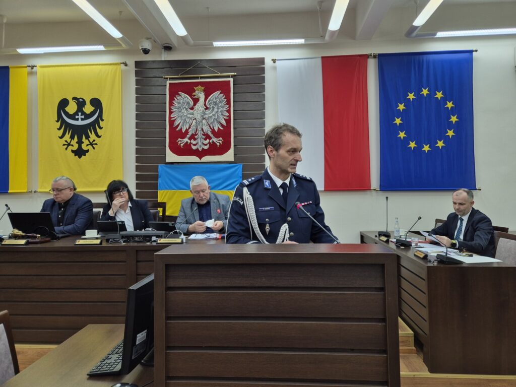 zastępca komendanta przemawia z mównicy, w tle prezydium rady