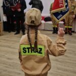dziewczynka w stroju strażackim stoi tyłem, w tle sztandar