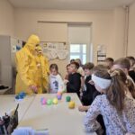 pracownicy sanepidu w żółtych kombinezonach z dziećmi, na stole kolorowe kulki