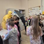 pracownicy sanepidu w żółtych kombinezonach, jeden trzyma parasol, grupa dzieci