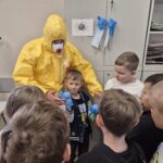 pracownik sanepidu w żółtym kombinezonie z grupa dzieci, chłopiec ubiera gumową rękawiczkę