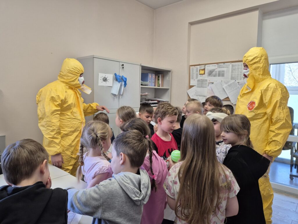 pracownicy sanepidu w żółtych kombinezonach z grupą dzieci