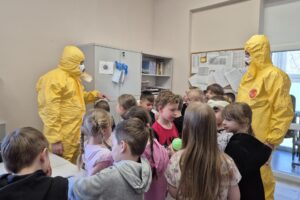pracownicy sanepidu w żółtych kombinezonach z grupą dzieci
