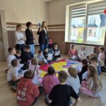 dzieci siedzą wokół planszy z grą, pracownice sanepidu stoją nad nimi