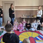 dzieci siedzą wokół planszy z grą, pracownice sanepidu stoją nad nimi