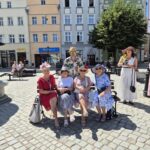 4 kobiety w kapeluszach na ławce, z tyłu wicestarosta, obok wicemarszałek